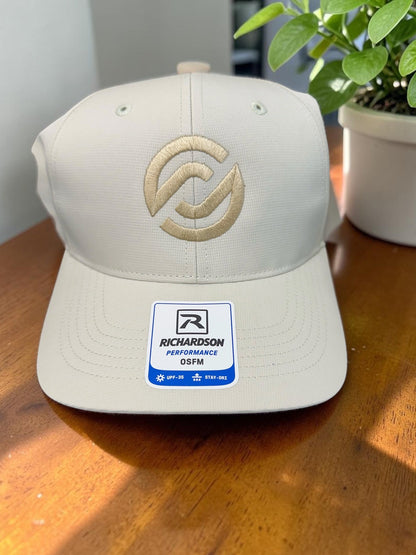 A light beige Richardson Performance 112PF baseball cap with a gold embroidered logo on the front, resting on a wooden table with a potted plant in the background.