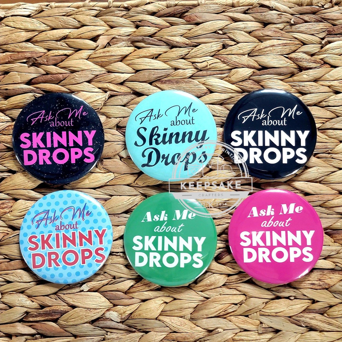 A collection of colorful pins with the text 'Ask Me About Skinny Drops' displayed on them, placed on a woven straw surface.