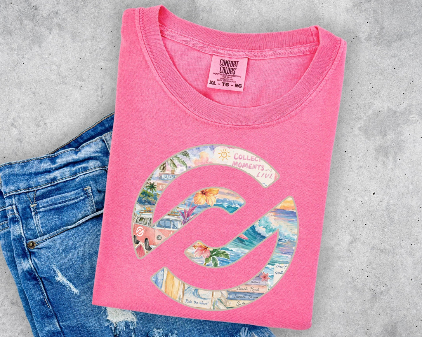 A folded pink t-shirt with a colorful graphic print, styled on top of a pair of blue jeans against a gray concrete background.