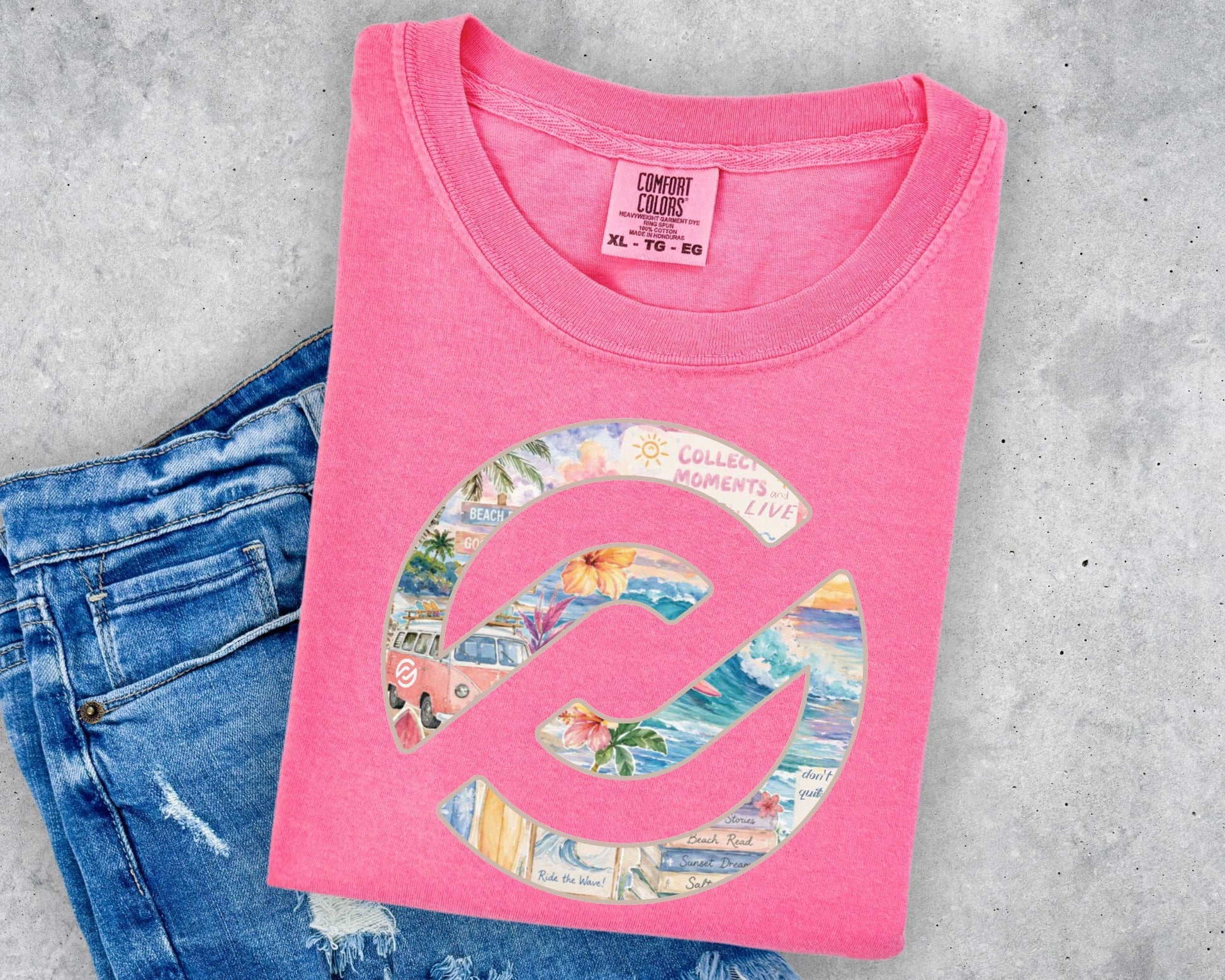 A folded pink t-shirt with a colorful graphic print, styled on top of a pair of blue jeans against a gray concrete background.