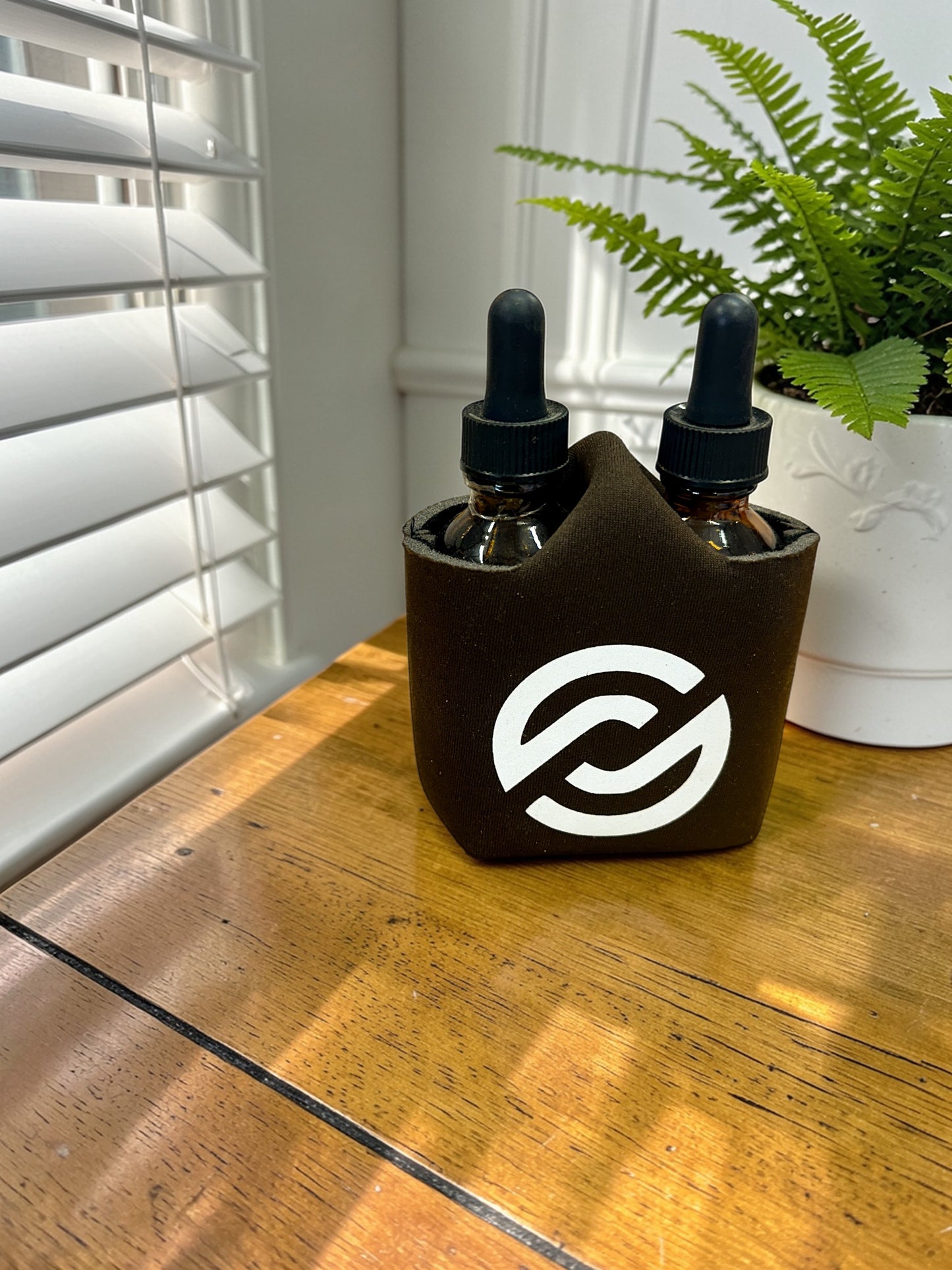 Two dropper bottles in a black sleeve with a partnerco logo on a wooden surface, next to a plant.