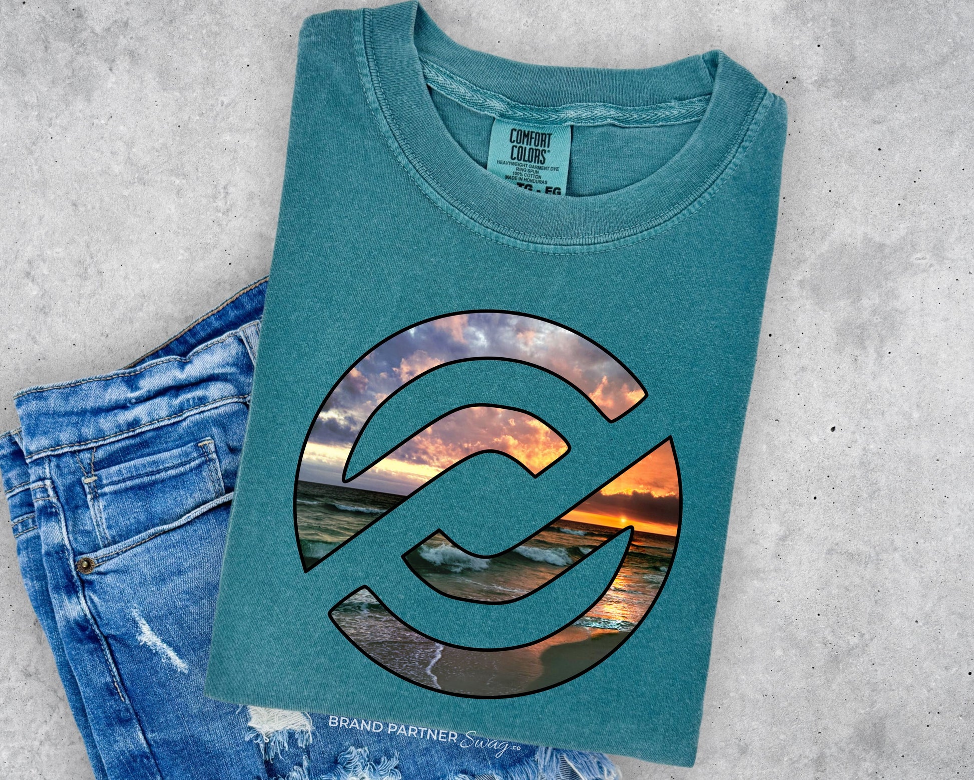 A folded teal t-shirt with a large circular graphic showing a sunset beach scene, styled flat-lay next to a pair of blue jeans on a gray concrete background.