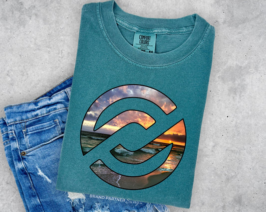 A folded teal t-shirt with a large circular graphic showing a sunset beach scene, styled flat-lay next to a pair of blue jeans on a gray concrete background.