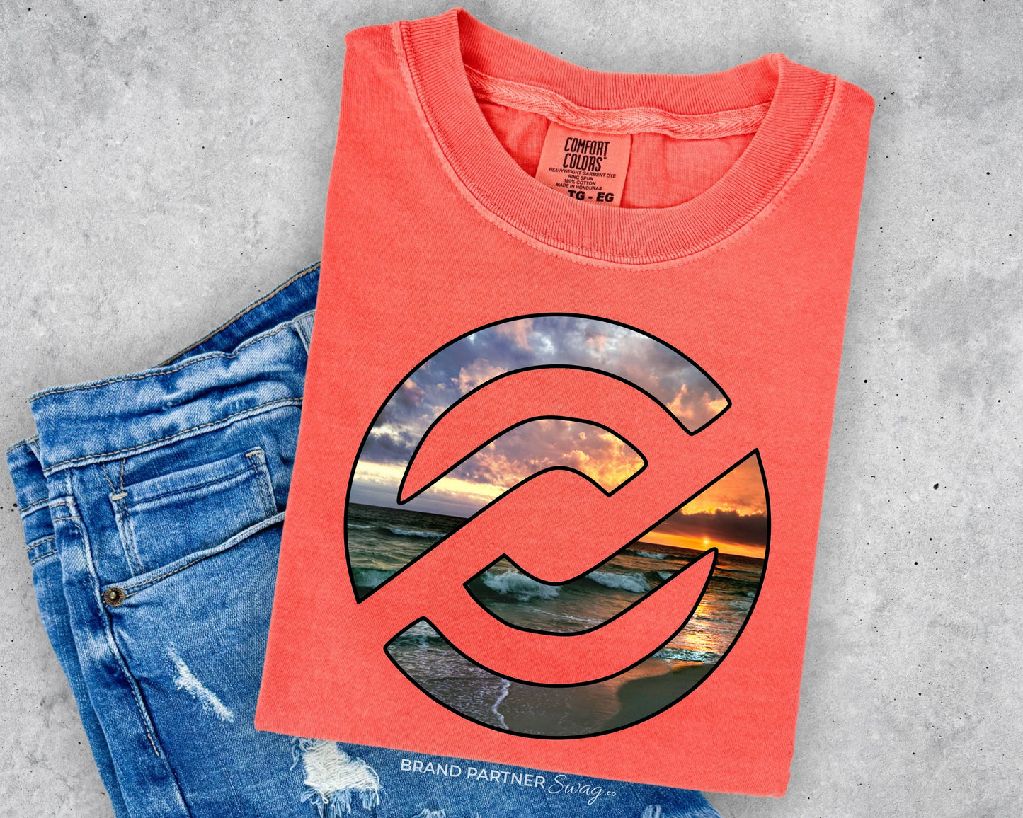 A coral pink t-shirt with a large circular graphic of a beach sunset, styled flat-lay on top of distressed blue jeans against a gray concrete background.