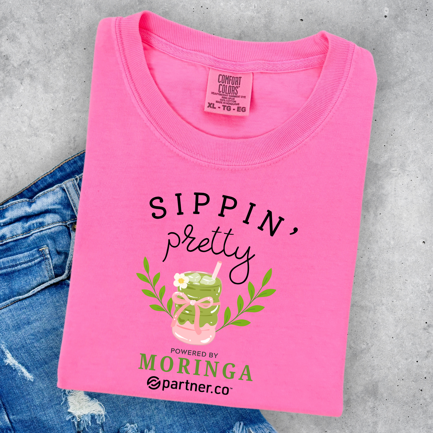 Pink Comfort Colors® graphic t-shirt featuring the phrase "Sippin' Pretty" with a Moringa tea drink illustration and feminine boutique-style design.