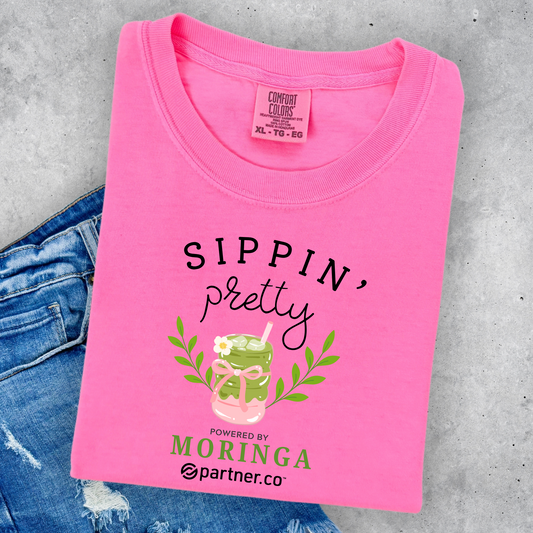 Pink Comfort Colors® graphic t-shirt featuring the phrase "Sippin' Pretty" with a Moringa tea drink illustration and feminine boutique-style design.
