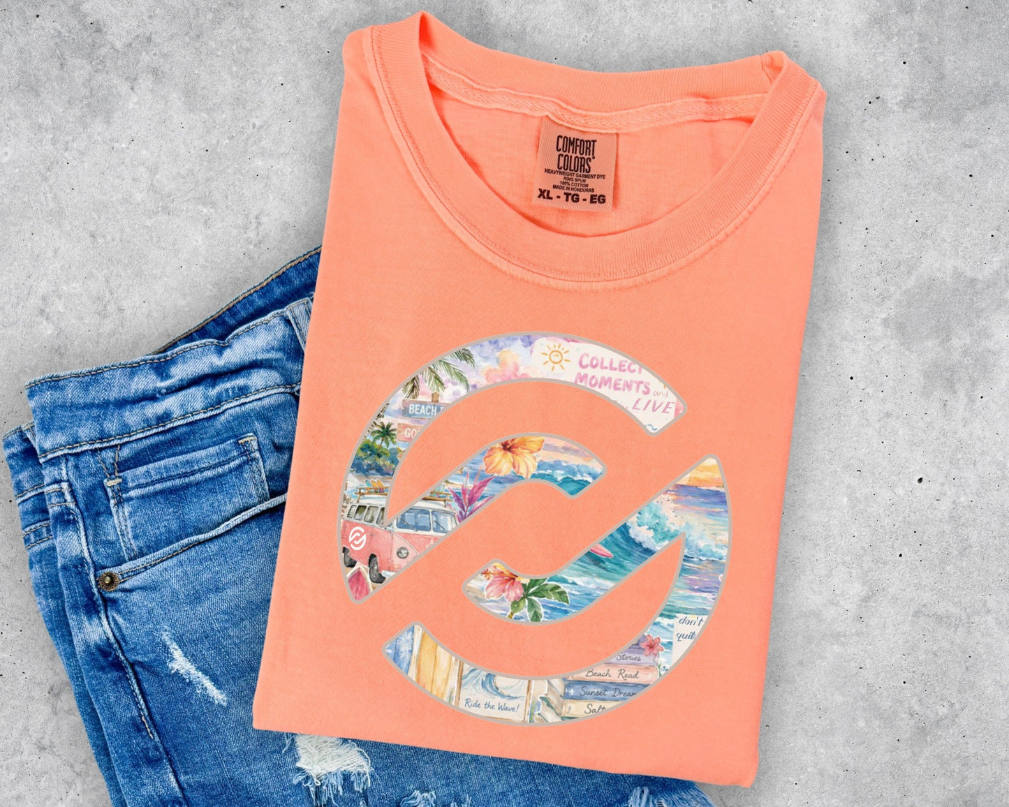 A folded coral orange t-shirt with a colorful graphic print, styled on top of a pair of blue jeans against a gray concrete background.