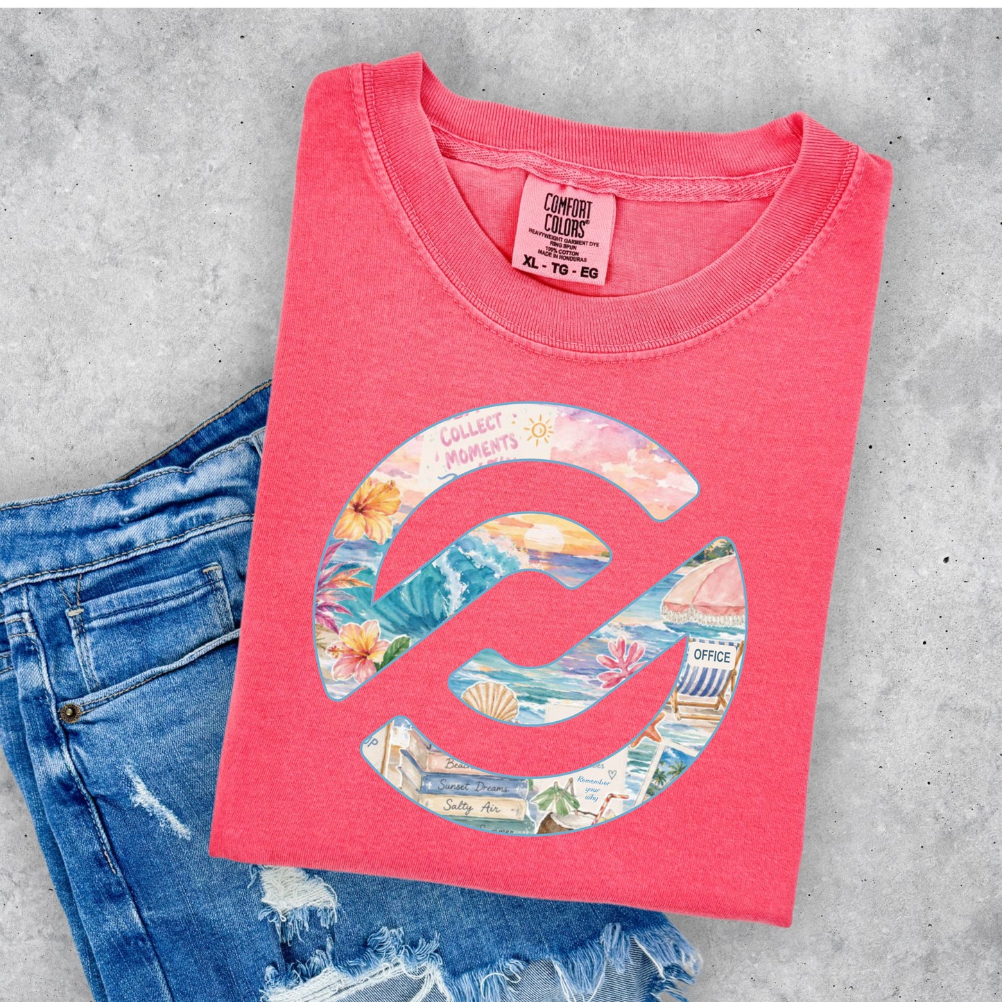A folded coral pink t-shirt with a colorful beach-themed circular graphic, styled on a gray concrete background next to a pair of distressed blue jeans. The neck label shows the brand Comfort Colors.