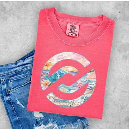 A folded coral pink t-shirt with a colorful beach-themed circular graphic, styled on a gray concrete background next to a pair of distressed blue jeans. The neck label shows the brand Comfort Colors.