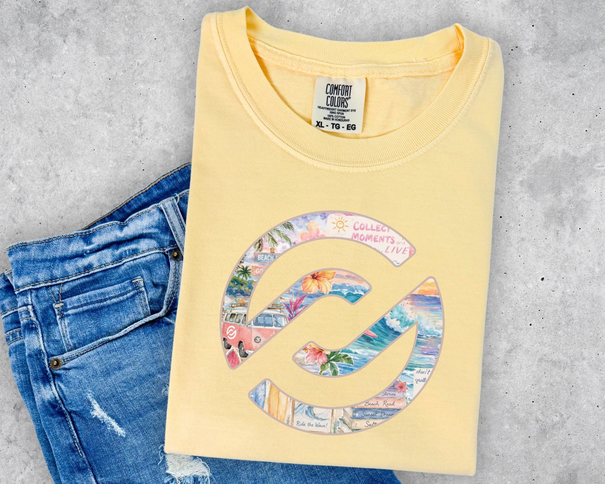 A folded pale yellow t-shirt with a colorful graphic print, styled flat-lay on top of a pair of blue denim shorts against a gray concrete background.