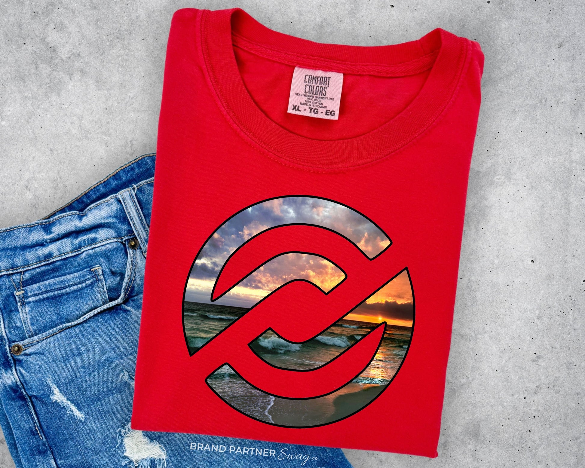 A folded red t-shirt with a large circular graphic on the front, styled flat-lay next to a pair of blue jeans on a gray concrete background. The graphic features a sunset beach scene with a red prohibition symbol overlaid.