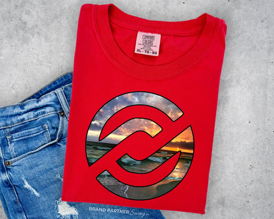 A folded red t-shirt with a large circular graphic on the front, styled flat-lay next to a pair of blue jeans on a gray concrete background. The graphic features a sunset beach scene with a red prohibition symbol overlaid.
