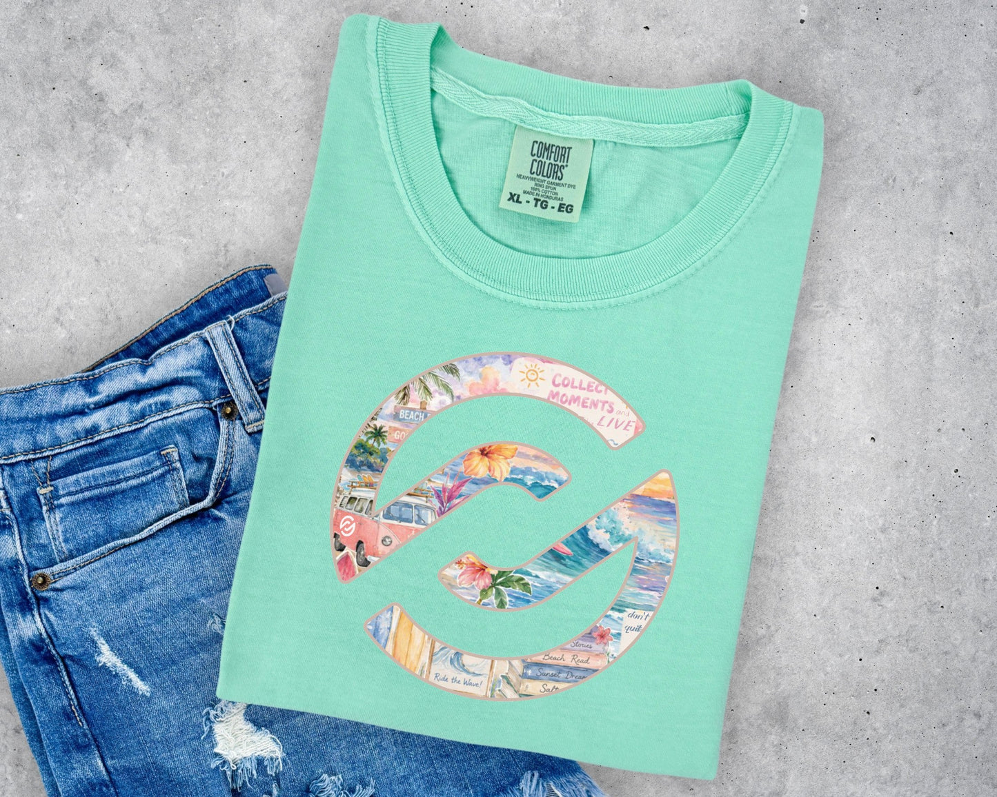 A mint green t-shirt with a colorful beach-themed graphic print, styled flat-lay on a gray concrete background next to a pair of blue jeans.
