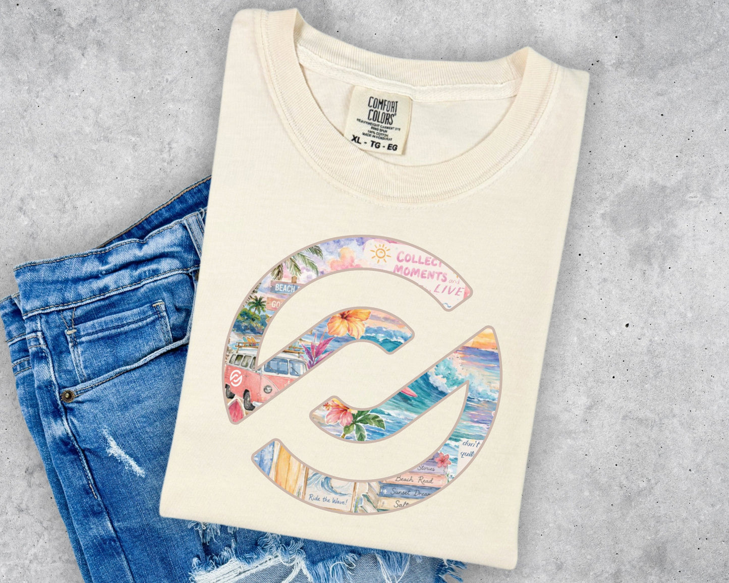 A cream-colored t-shirt with a colorful graphic of a spiral shell containing beach scenes and the text 'COLLECTED MOMENTS COCO', styled flat-lay on blue jeans against a gray concrete background.