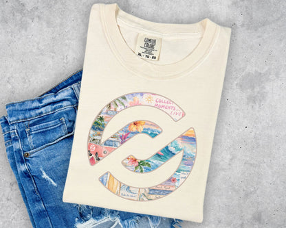 A cream-colored t-shirt with a colorful graphic of a spiral shell containing beach scenes and the text 'COLLECTED MOMENTS COCO', styled flat-lay on blue jeans against a gray concrete background.