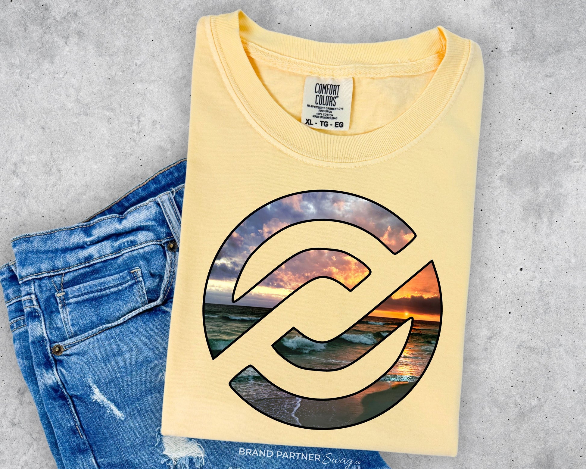 A folded pale yellow t-shirt with a large circular graphic of a beach sunset, styled flat-lay next to a pair of blue jeans on a gray concrete background. The neck label shows the brand 'Comfort Colors'.
