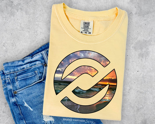 A folded pale yellow t-shirt with a large circular graphic of a beach sunset, styled flat-lay next to a pair of blue jeans on a gray concrete background. The neck label shows the brand 'Comfort Colors'.