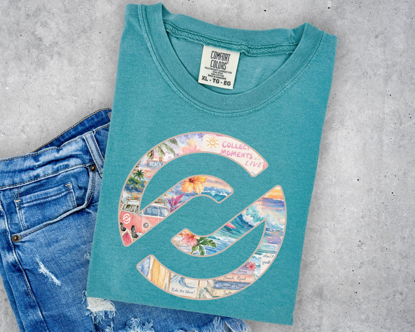 A teal t-shirt with a colorful graphic print, styled flat-lay on a gray concrete background next to a pair of blue jeans. The shirt features a large circular 'C' logo filled with a collage of beach and travel images, with the text 'COLLECTED MOMENTS' arched over the top.
