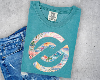 A teal t-shirt with a colorful graphic print, styled flat-lay on a gray concrete background next to a pair of blue jeans. The shirt features a large circular 'C' logo filled with a collage of beach and travel images, with the text 'COLLECTED MOMENTS' arched over the top.