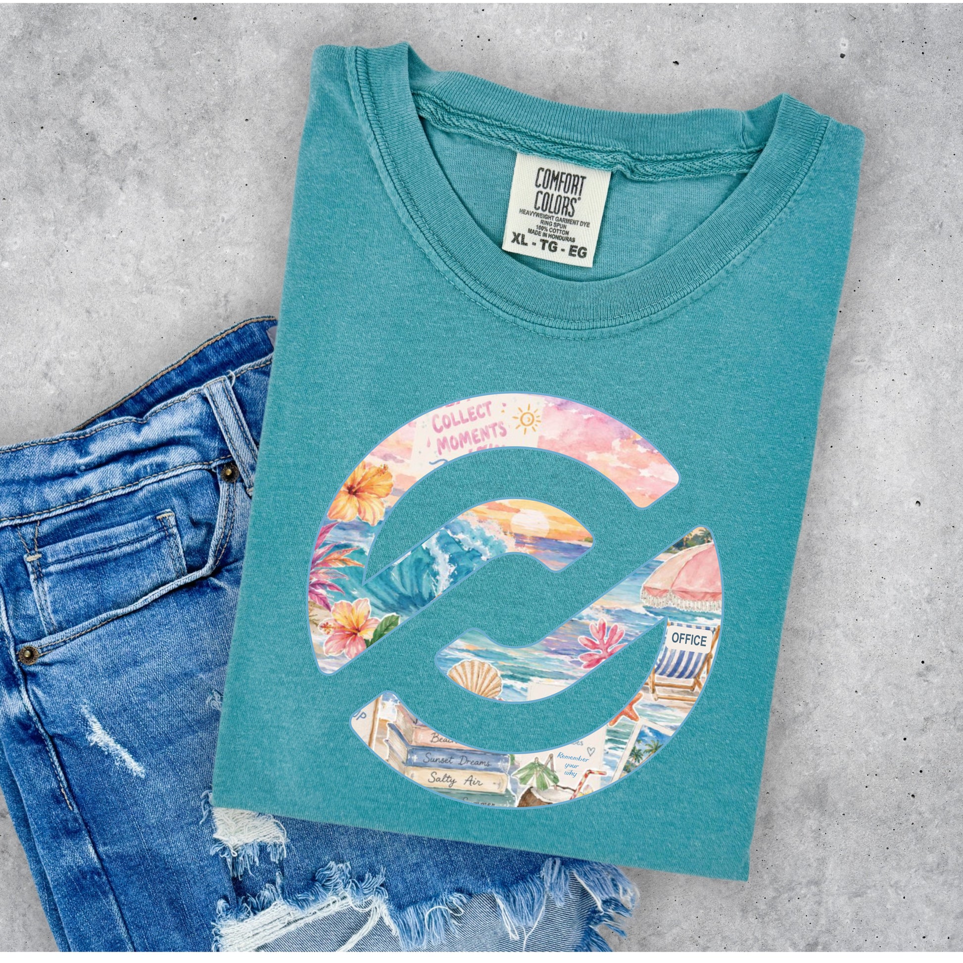 A teal t-shirt with a colorful graphic print, styled flat-lay on a gray concrete background next to a pair of blue jeans. The shirt features a large llogo filled with a collage of beach and travel images, with the text 'COLLECT MOMENTS' arched over the top.