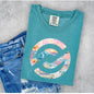 A teal t-shirt with a colorful graphic print, styled flat-lay on a gray concrete background next to a pair of blue jeans. The shirt features a large llogo filled with a collage of beach and travel images, with the text 'COLLECT MOMENTS' arched over the top.
