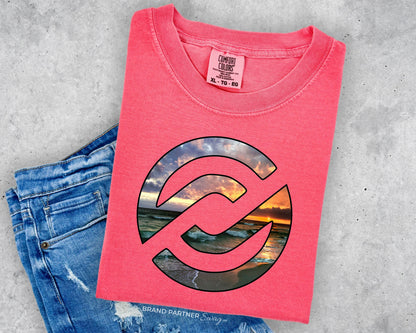 A folded coral pink t-shirt with a circular graphic of a sunset over water, styled on top of blue denim jeans against a gray concrete background.