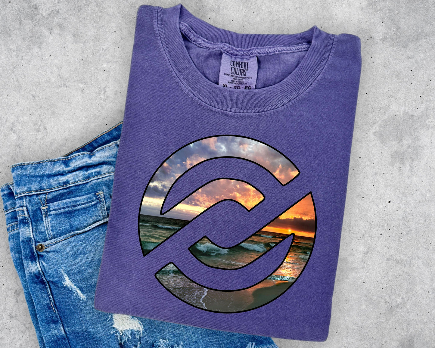 A folded purple t-shirt with a circular graphic of a sunset beach scene, styled on top of a pair of blue denim jeans against a gray concrete background.
