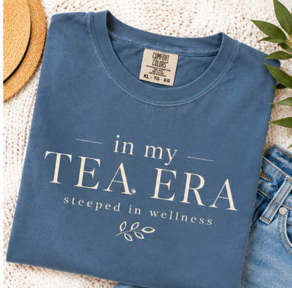 Comfort Colors Blue Jean t-shirt featuring the phrase "In My Tea Era" with "Steeped in Wellness" typography and a minimalist botanical design inspired by coastal boutique style.