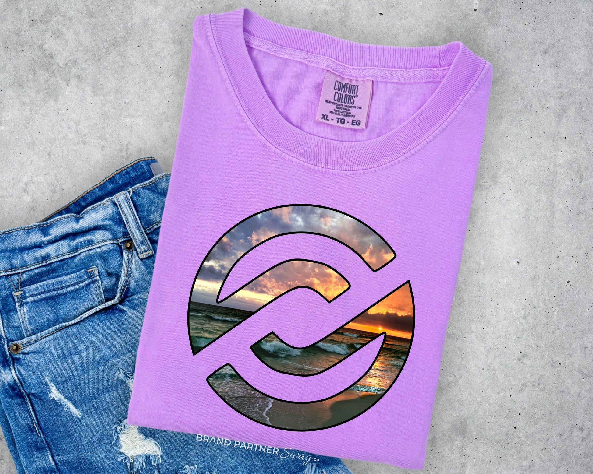 A folded lavender-purple t-shirt with a circular graphic of a sunset over water, styled on top of a pair of blue jeans against a gray concrete background.