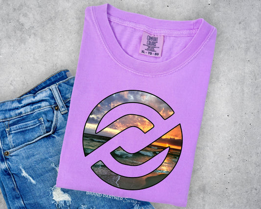 A folded lavender-purple t-shirt with a circular graphic of a sunset over water, styled on top of a pair of blue jeans against a gray concrete background.